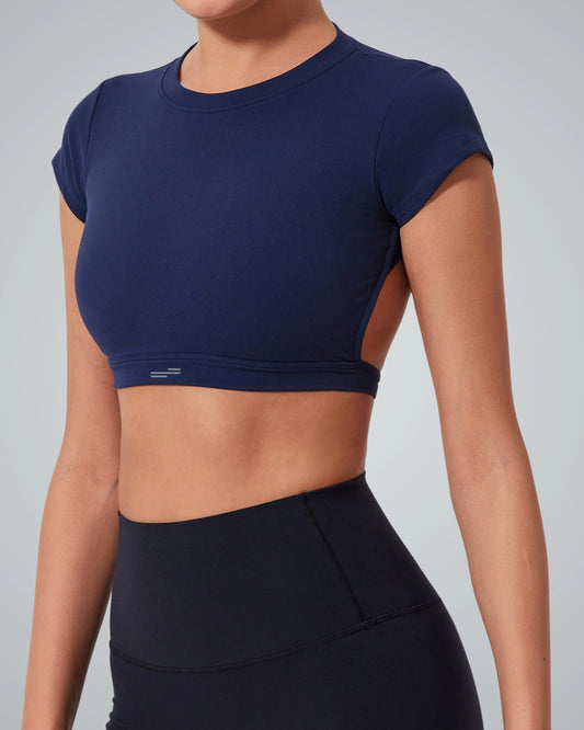 A woman wearing a Bandsam fitted short-sleeve navy ScarletFlow Top with an open back and high-waisted navy leggings stands against a light gray background. Her face is partially out of frame, showcasing a chic exercise wardrobe.