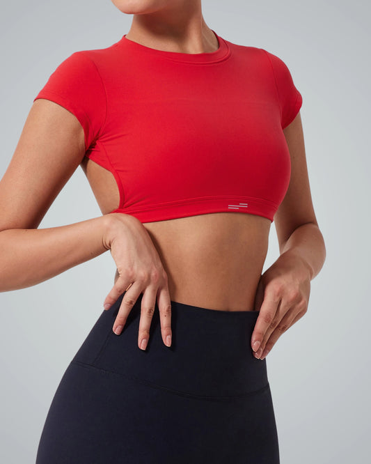 A woman wearing a Bandsam ScarletFlow Top—an open back, red short-sleeve crop ideal for your exercise wardrobe—and high-waisted black leggings poses with one hand on her hip and the other resting on her waist, against a plain light background.