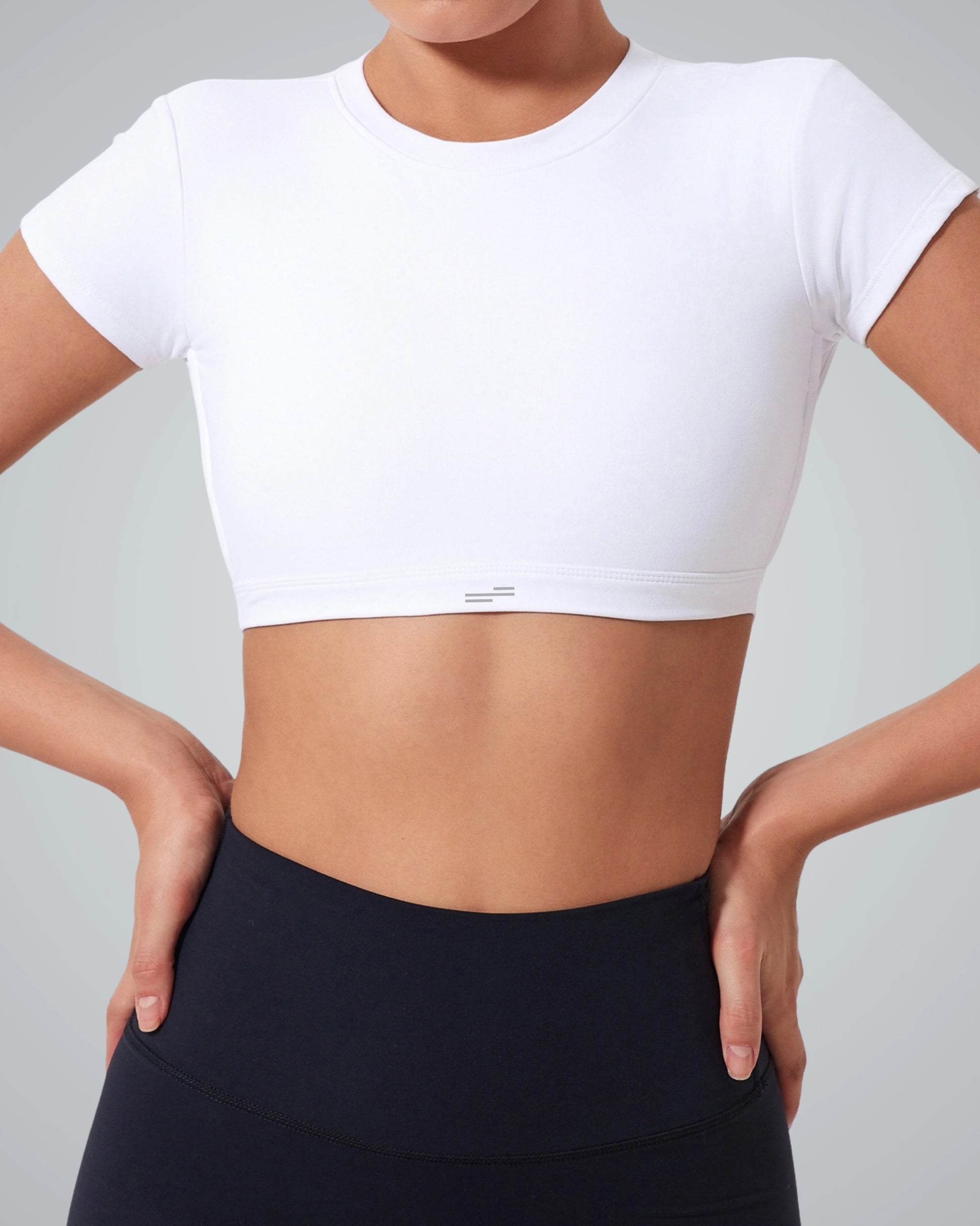 A woman wearing a Bandsam fitted white ScarletFlow Top with an open back and high-waisted black leggings stands with her hands on her hips against a plain gray background, showcasing her exercise wardrobe.