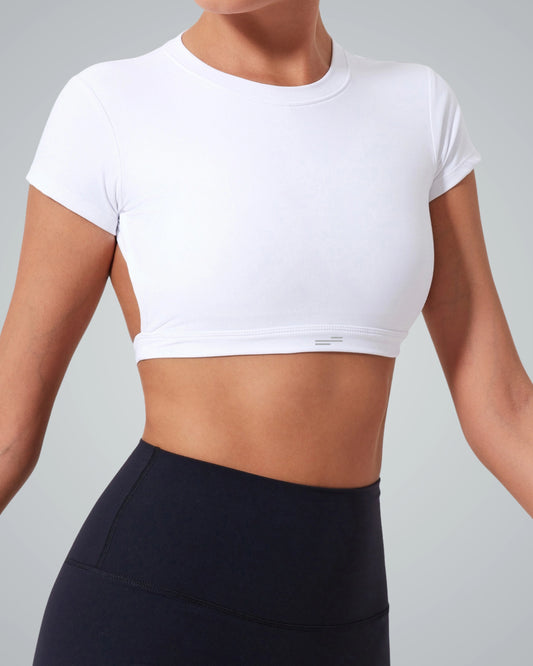 A woman wears a Bandsam white open back top with short sleeves and high-waisted black leggings, standing against a light gray background—perfect for elevating your exercise wardrobe.